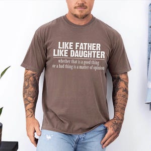 May include: Brown t-shirt with the text "LIKE FATHER LIKE DAUGHTER" in white, with the phrase "whether that is a good thing or a bad thing is a matter of opinion" underneath. The shirt is worn by a person with tattoos on their arms.