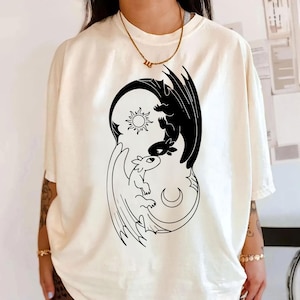 May include: Cream-colored t-shirt featuring a black and white dragon design. The graphic depicts two dragons in a yin-yang style, with a sun and moon. The shirt has short sleeves and a relaxed fit.