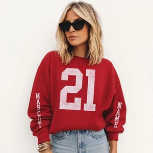 A woman wearing a red sweatshirt with the number 21 on it, sunglasses, and jeans stands against a white background.