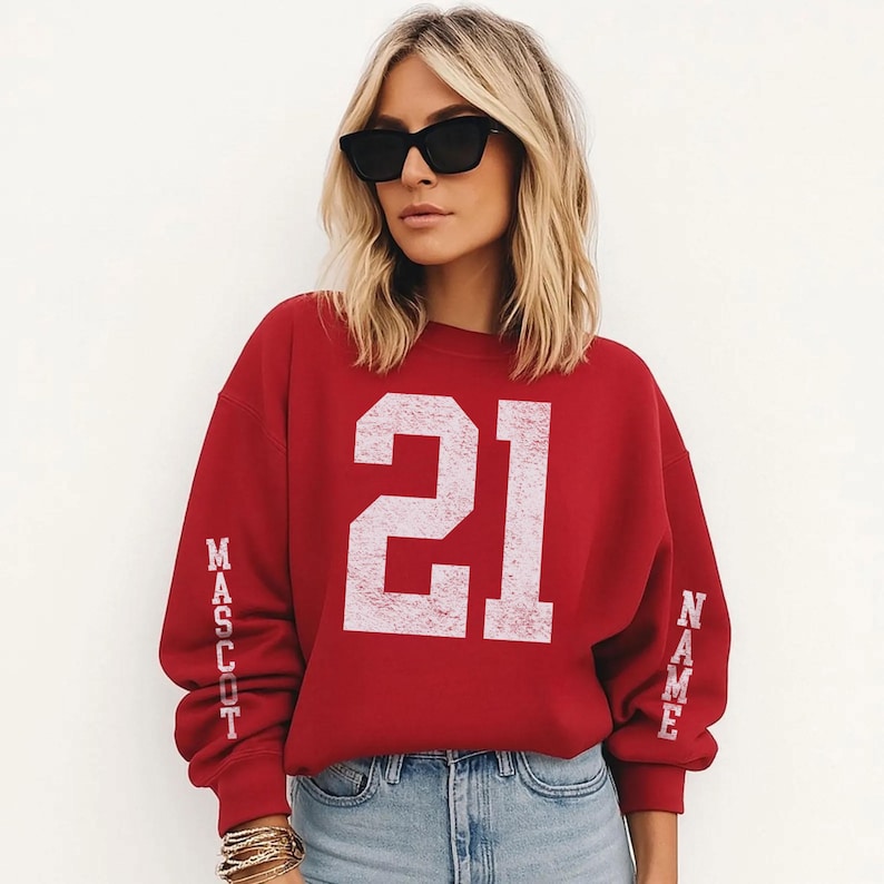 A woman wearing a red sweatshirt with the number 21 on it, sunglasses, and jeans stands against a white background.