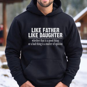 May include: Black hooded sweatshirt with the text "LIKE FATHER LIKE DAUGHTER" in white, with the phrase "whether that is a good thing or a bad thing is a matter of opinion" underneath. The sweatshirt is a solid black color.
