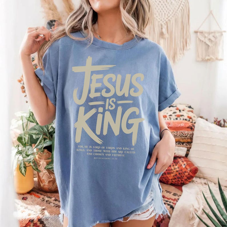 A woman wearing a blue t-shirt with the text "JESUS IS KING" printed on it, standing in a room with various decorative elements such as plants and a woven wall hanging.