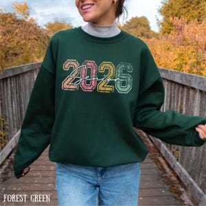 A woman wearing a green sweatshirt with the text "2026" printed on it, standing on a wooden bridge surrounded by autumn foliage.