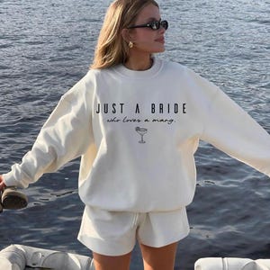 May include: Cream-colored sweatshirt and shorts set. The sweatshirt features the text "JUST A BRIDE who loves a marg." with a margarita glass graphic. The outfit is worn by a person with sunglasses and gold earrings.