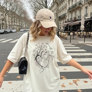 May include: Cream-colored t-shirt with a black and white anatomical heart design, surrounded by flowers and the text "Go with the flow." A beige baseball cap with a small black logo is worn.