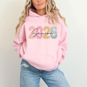 A woman wearing a pink hoodie with the text "2026" printed on it.