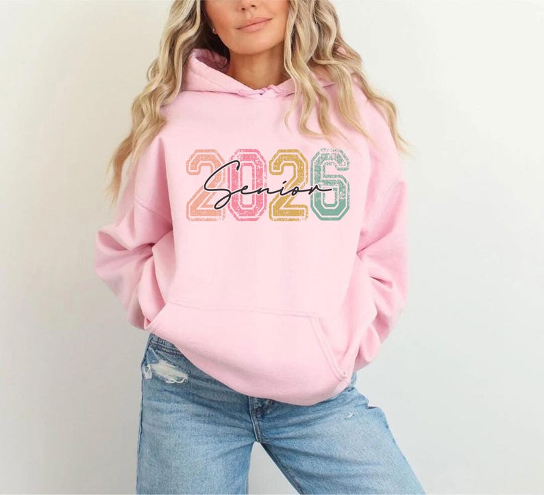 A woman wearing a pink hoodie with the text "2026" printed on it.