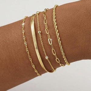 Può includere: Una collezione di cinque braccialetti dorati, ognuno con un design unico. I braccialetti includono uno stile a corda intrecciata, un motivo a spina di pesce, una catena con piccole perle, una catena con maglie ovali e una delicata catena intrecciata.