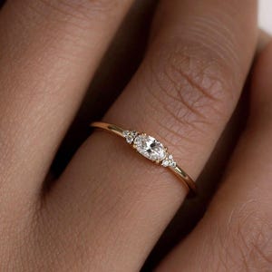 May include: A delicate gold ring featuring an oval-cut diamond center stone, flanked by smaller accent diamonds. The ring's band is thin and polished, creating a minimalist and elegant design. The ring is shown on a finger, highlighting its fine details and sparkle.