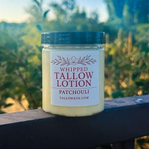 Puede incluir: Un tarro de loción de sebo batida con una etiqueta blanca. La etiqueta dice "Whipped Tallow Lotion Patchouli" con un diseño floral. El tarro tiene una tapa verde oscuro y está sobre un fondo verde borroso.