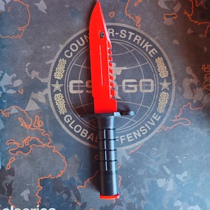 Pode incluir: Uma faca de brinquedo vermelha e preta com uma borda serrilhada, em um fundo com o texto "Counter-Strike: Global Offensive". A faca tem uma alça preta e uma lâmina vermelha brilhante.