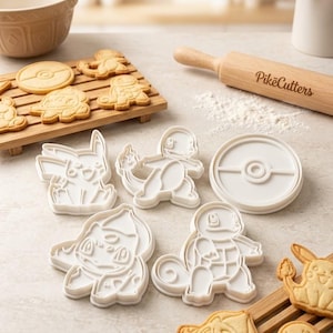 May include: A set of four white plastic cookie cutters featuring character shapes and a circular design, accompanied by a wooden rolling pin with the text PikeCutters engraved on it, alongside several baked cookies on wooden racks.