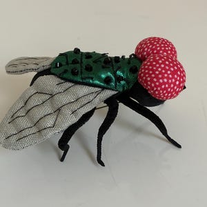 May include: A whimsical fly brooch with silver wings, black legs, and a green body adorned with black beads. The fly's eyes are two red spheres with white polka dots. This unique accessory adds a touch of playful charm.