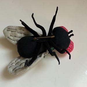 May include: A handmade fly brooch with a black body, legs, and antennae. The wings are silver with a textured pattern. The fly's head is red with white polka dots. A gold-colored pin is attached to the back.