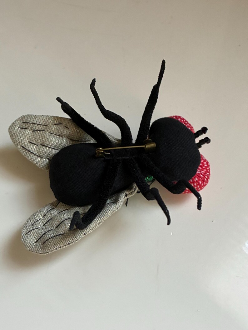May include: A handmade fly brooch with a black body, legs, and antennae. The wings are silver with a textured pattern. The fly's head is red with white polka dots. A gold-colored pin is attached to the back.