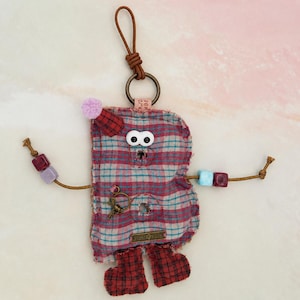 May include: A handmade fabric keychain shaped like the letter "B", featuring a plaid pattern in red, blue, and white. It has googly eyes, beaded arms, and small red plaid boots. A brown leather loop and metal ring are attached for hanging.