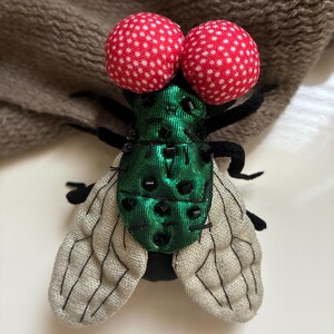 May include: A decorative fly pin with large red polka-dot eyes. The fly's body is green with black bead accents, and it has gray and black wings. The pin is made of fabric and is likely handmade.