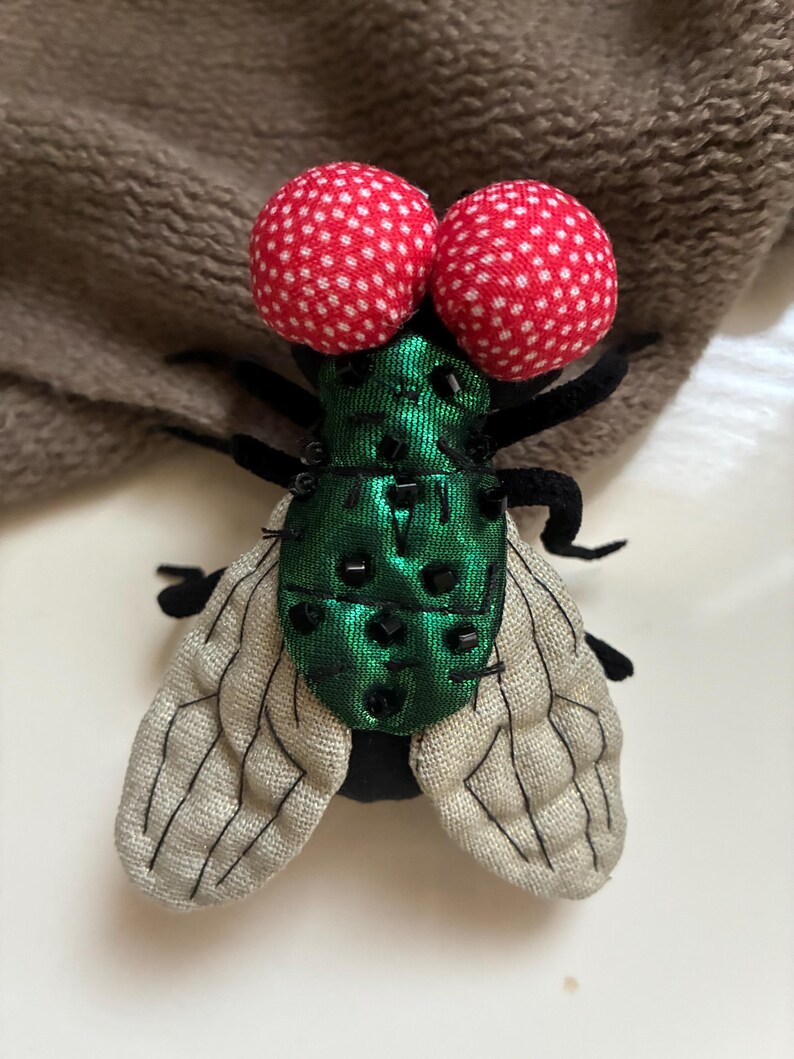 May include: A decorative fly pin with large red polka-dot eyes. The fly's body is green with black bead accents, and it has gray and black wings. The pin is made of fabric and is likely handmade.