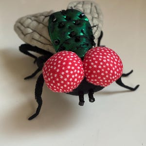 May include: A whimsical, handmade fly figurine. The fly features large, red polka-dotted eyes, a green iridescent body, and gray wings. Its legs and antennae are black. This unique piece could be used as a decorative item or a quirky accessory.