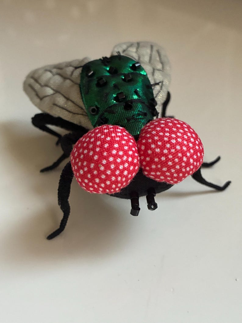 May include: A whimsical, handmade fly figurine. The fly features large, red polka-dotted eyes, a green iridescent body, and gray wings. Its legs and antennae are black. This unique piece could be used as a decorative item or a quirky accessory.