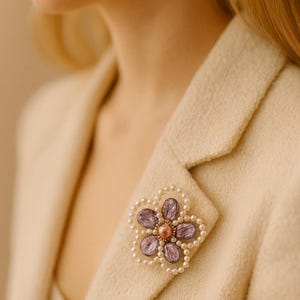 May include: A floral brooch with a pearl border and purple petals is pinned to a cream-colored blazer. The brooch's center features a small, round, copper-colored bead. The blazer's fabric has a textured appearance.
