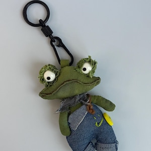 May include: A green frog keyring with large white eyes and denim dungarees. The frog has a black metal keyring and clasp. A yellow button with a smiling face is on the dungarees.