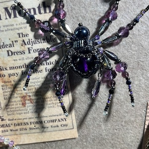 May include: A beaded spider ornament with purple, black, and clear beads. The spider has multiple legs and a detailed body. The background includes a vintage advertisement with text.