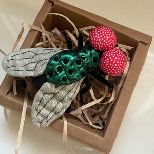 May include: A handcrafted fly brooch with a green, textured body, gray wings, and two red, polka-dotted eyes. The insect-themed accessory is presented in a small, brown cardboard box with shredded paper filler.