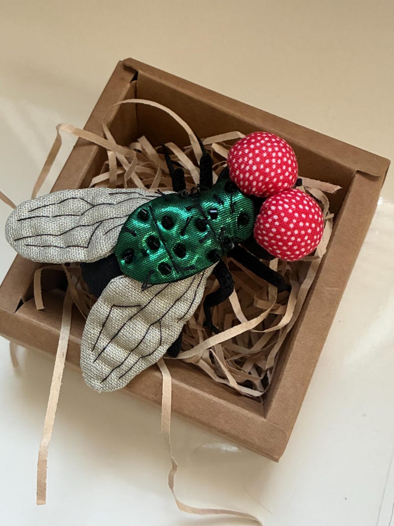 May include: A handcrafted fly brooch with a green, textured body, gray wings, and two red, polka-dotted eyes. The insect-themed accessory is presented in a small, brown cardboard box with shredded paper filler.