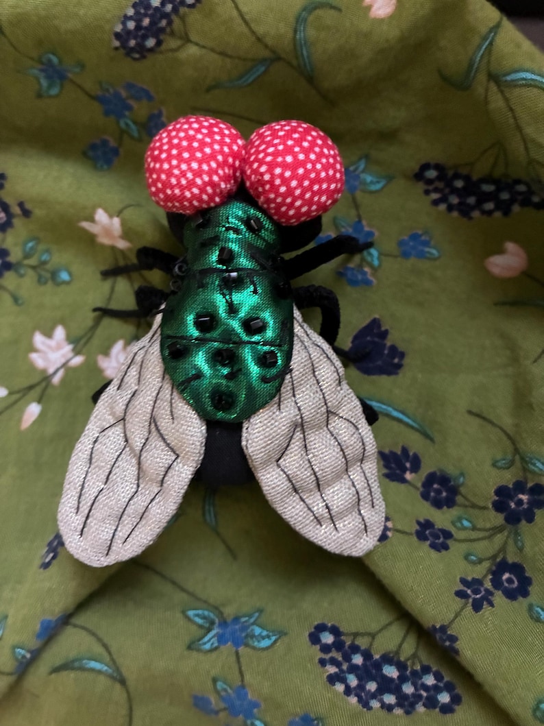 May include: A whimsical fly brooch with large red and white polka-dotted eyes, a shimmering green body, and delicate, patterned wings. The insect-themed accessory is set against a green floral fabric.