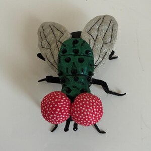 May include: A whimsical fly-shaped brooch. The body is green with black accents, and the wings are a light gray fabric. Two red, polka-dotted spheres form the lower portion of the fly's body. The brooch has black legs and antennae.