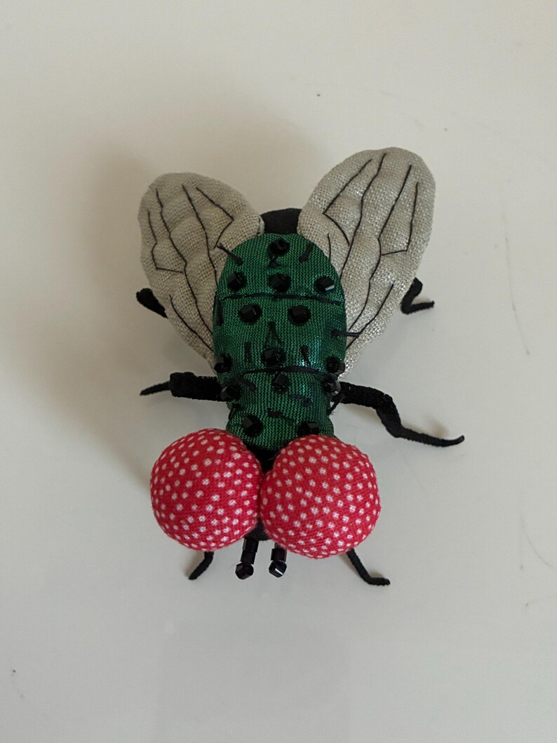 May include: A whimsical fly-shaped brooch. The body is green with black accents, and the wings are a light gray fabric. Two red, polka-dotted spheres form the lower portion of the fly's body. The brooch has black legs and antennae.