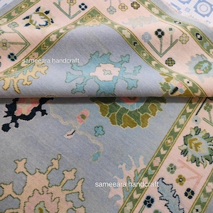 May include: A close-up of a light blue rug with detailed floral and geometric patterns in pink, green, and cream. The rug has a border design and the text "sameeara handcraft".