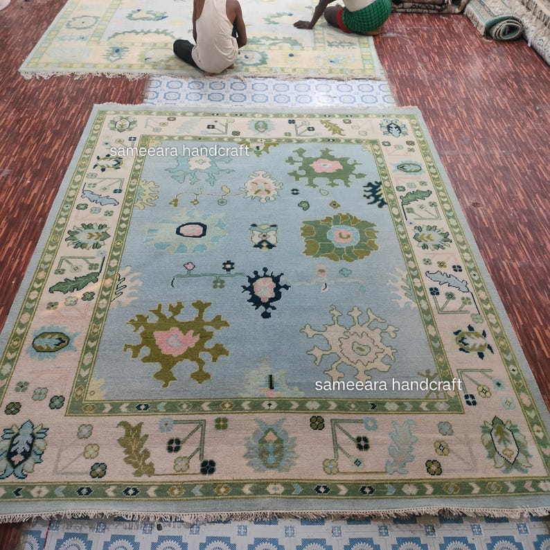 May include: A large, light blue rug with a detailed floral pattern and a border of green and beige. The rug features various floral and geometric designs in shades of green, pink, and white. The text "sameeara handcraft" is visible on the rug.