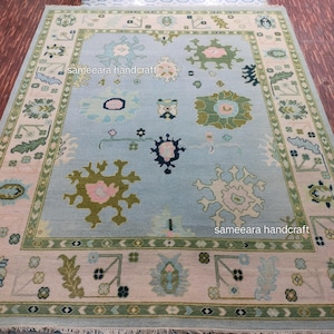 May include: A large, rectangular rug with a light blue central field. The rug features a repeating pattern of floral and geometric designs in shades of green, pink, and cream. The border is a contrasting cream colour with green accents. The rug has the text "sameeara handcraft".