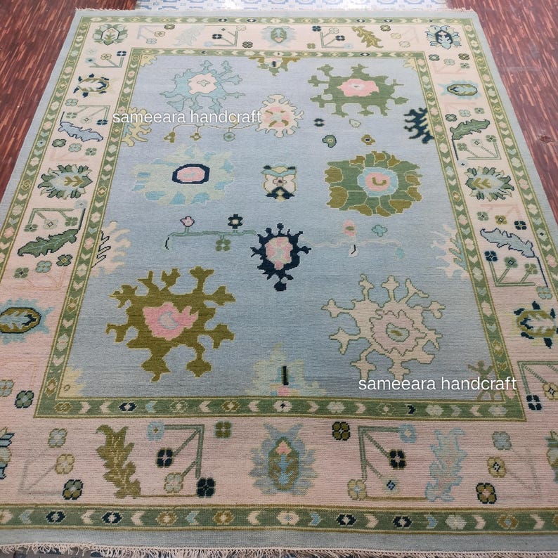 May include: A large, rectangular rug with a light blue central field. The rug features a repeating pattern of floral and geometric designs in shades of green, pink, and cream. The border is a contrasting cream colour with green accents. The rug has the text "sameeara handcraft".