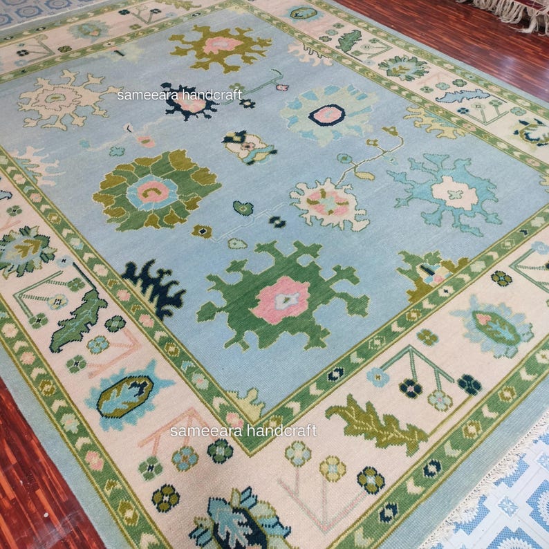 May include: A large, rectangular rug with a light blue central field and a cream-coloured border. The rug features a repeating floral pattern in shades of green, pink, and blue. The border is accented with a green geometric design. The words "sameeara handcraft" are visible.
