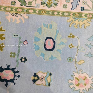 May include: A close-up of a light blue rug with floral patterns. The rug features large, stylized flowers in shades of blue, pink, and green. The border has a cream background with green and pink accents. The text "sameeara handcraft" is visible.