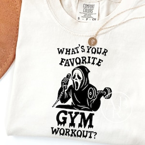 May include: Cream-colored t-shirt with the text "WHAT'S YOUR FAVORITE GYM WORKOUT?" and a black graphic of a ghost holding a dumbbell and a phone. The shirt has a crew neck and short sleeves. A gold necklace is draped over the shirt.