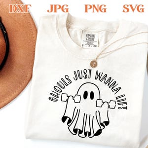 May include: Cream-colored t-shirt with the phrase "Ghouls Just Wanna Lift" in black. The design features a ghost lifting weights. The shirt has a round neckline and short sleeves. A gold necklace with a pendant is draped over the shirt.
