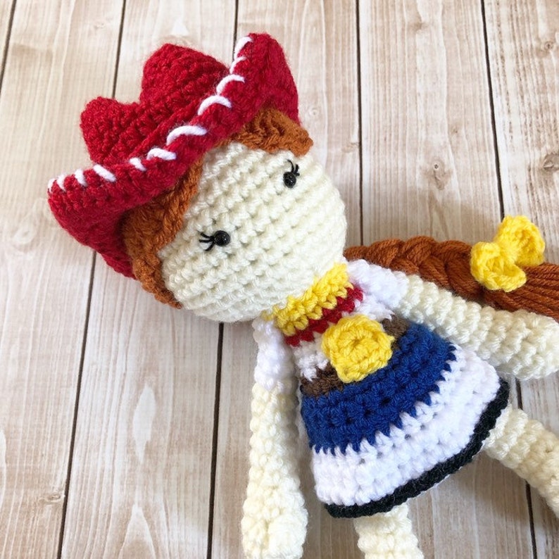 May include: Crocheted doll with a red cowboy hat, brown hair, and a blue and white dress. The doll is wearing a yellow sheriff's star.