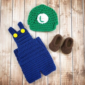 May include: Crocheted baby outfit in blue and green. The outfit includes overalls, a hat, and shoes. The hat has a green and white L on it.