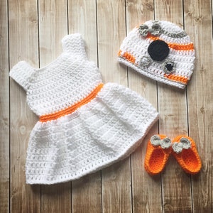 May include: A white crocheted dress with an orange belt, a white and orange crocheted hat with a black circle and gray details, and a pair of orange crocheted booties with gray flowers.