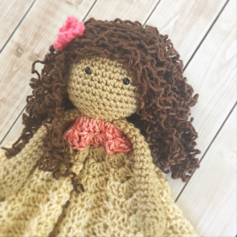 Pode incluir: Uma boneca de croch&ecirc; com cabelo encaracolado castanho, uma flor rosa e um vestido de croch&ecirc; cor de p&ecirc;ssego. A boneca est&aacute; deitada sobre uma superf&iacute;cie de madeira marrom claro.