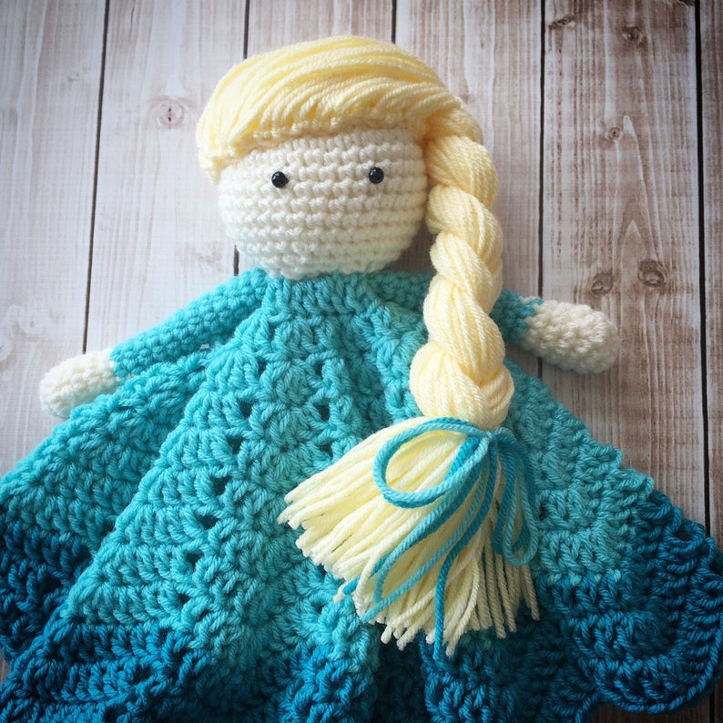 May include: A crocheted doll with a long blonde braid, wearing a blue and teal crocheted dress. The doll is holding a blue and teal crocheted blanket.