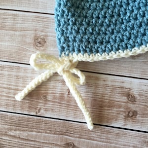 May include: A blue crocheted baby hat with a white crocheted brim and matching ties. The hat is tied with a bow.