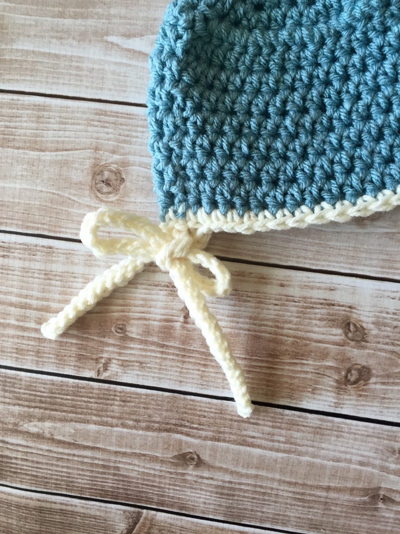 May include: A blue crocheted baby hat with a white crocheted brim and matching ties. The hat is tied with a bow.