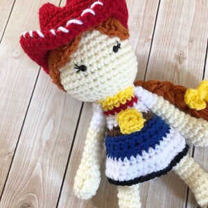 May include: Crocheted doll with a red and white cowboy hat, brown hair, and a blue and white dress with a yellow star.