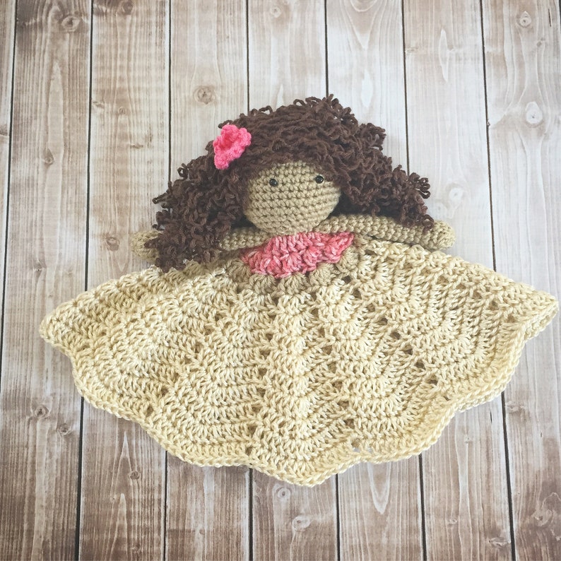 Pode incluir: Uma boneca de croch&ecirc; com cabelo castanho encaracolado e uma flor rosa. A boneca est&aacute; vestindo um vestido rosa e bege e est&aacute; deitada em um cobertor de croch&ecirc; bege.