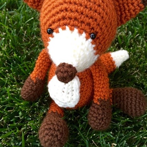 May include: A crocheted fox plush toy with brown, orange, and white yarn. The fox is sitting on a bed of green grass.
